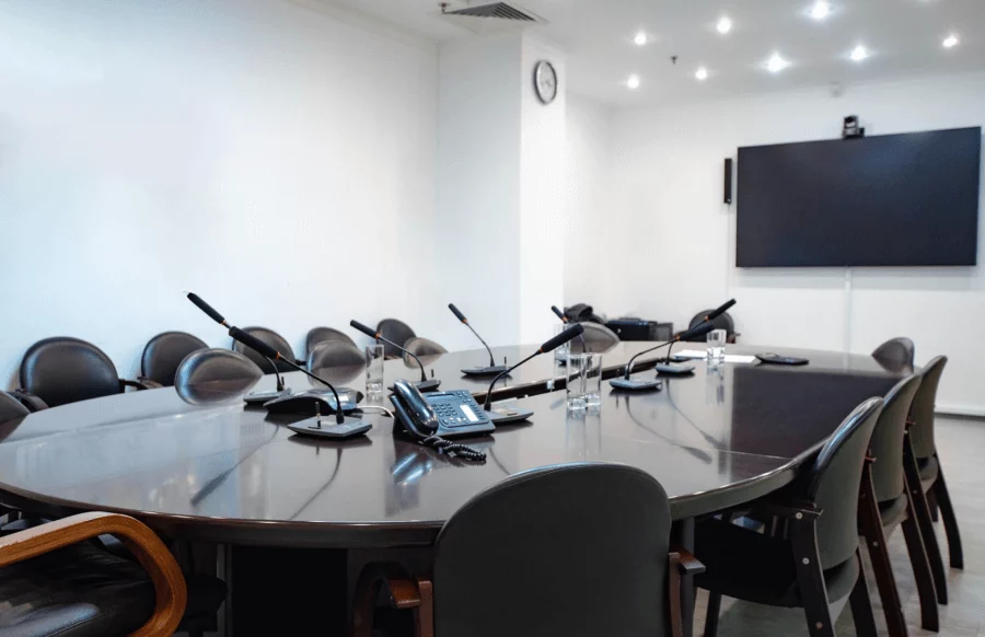 A conference room setting showcases the comparison of Huawei IdeaHub vs Traditional VC Systems. A large, oval wooden table is surrounded by multiple black chairs. Microphones are positioned on the table. A large screen, possibly a Huawei IdeaHub, is mounted on the wall.