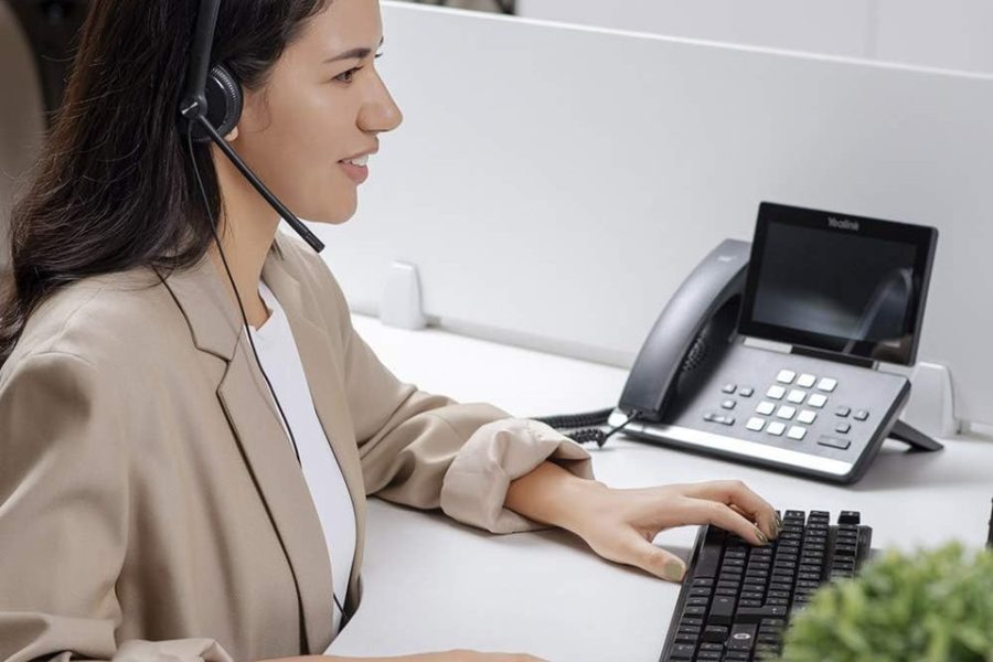 Smiling woman in a beige blazer using a wired headset while working on a computer at an office desk with a Yealink phone beside her.