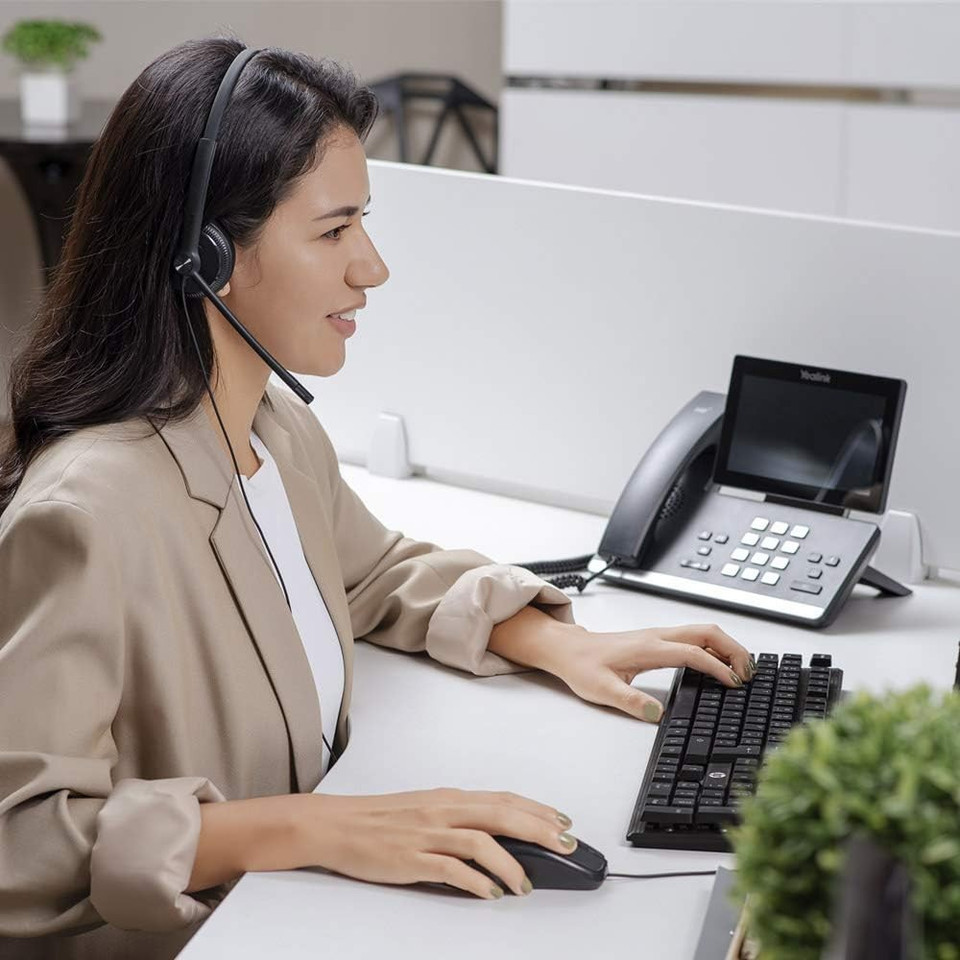 Smiling woman in a beige blazer using a wired headset while working on a computer at an office desk with a Yealink phone beside her.