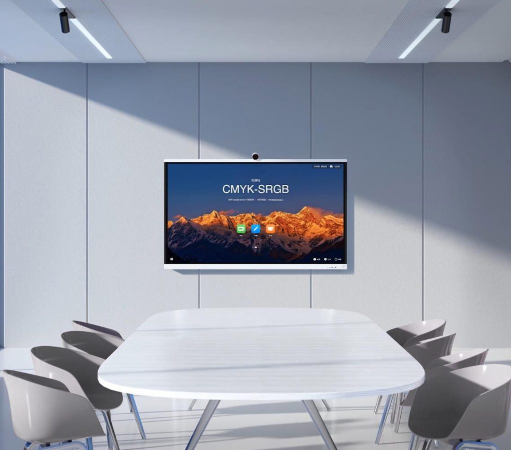 Essential Video Conferencing Upgrades for Large Meeting Rooms: A sleek, wall-mounted Huawei IdeaHub S2 dominates the view above a large, modern white conference table surrounded by grey chairs in a well-lit meeting space. The display shows a vibrant image of snow-capped mountains under a warm sky and the text "CMYK-SRGB," suggesting advanced color management capabilities for professional video conferencing.