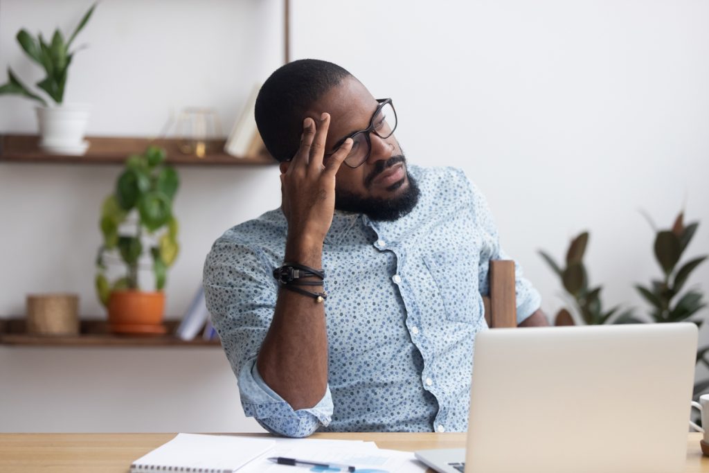 A Black man with a beard and glasses rests his head on his hand, looking away from an open laptop on a wooden desk, perhaps experiencing a common video conferencing problem. He appears tired or frustrated. In the background, there's a white wall with wooden shelves holding potted green plants. There are also papers and a pen on the desk.