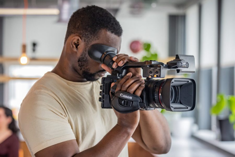 A man using a Canon XF605 professional 4K camcorder, looking through the viewfinder while filming indoors in a modern, well-lit space with blurred background elements.