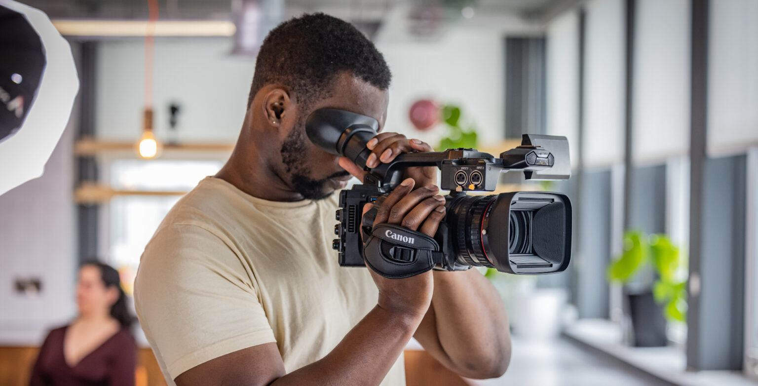 A man using a Canon XF605 professional 4K camcorder, looking through the viewfinder while filming indoors in a modern, well-lit space with blurred background elements.
