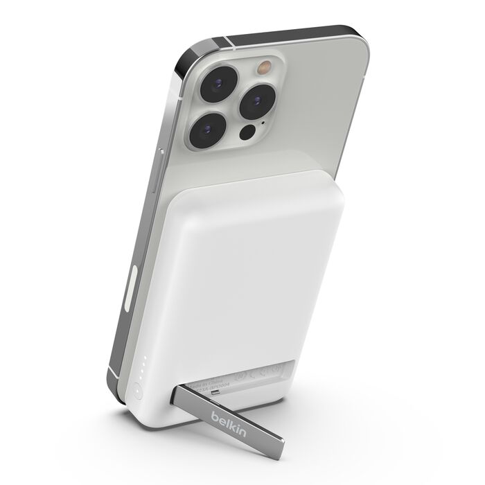 A white Belkin magnetic wireless power bank with a silver kickstand attached to the back of a silver iPhone. The power bank has rounded edges and an LED indicator with four lights visible on its bottom left side. The iPhone is slightly angled, showing its three camera lenses and the side button. The background is a solid white.