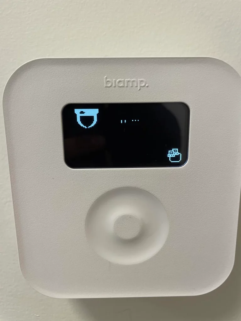 A close-up of a white Biamp Tesira TEC-1i control panel mounted on a wall. The black screen displays a pixelated bucket icon on the left, an ellipsis in the center, and a box icon with an arrow pointing out of it on the right. Below the screen is a circular, textured control dial.