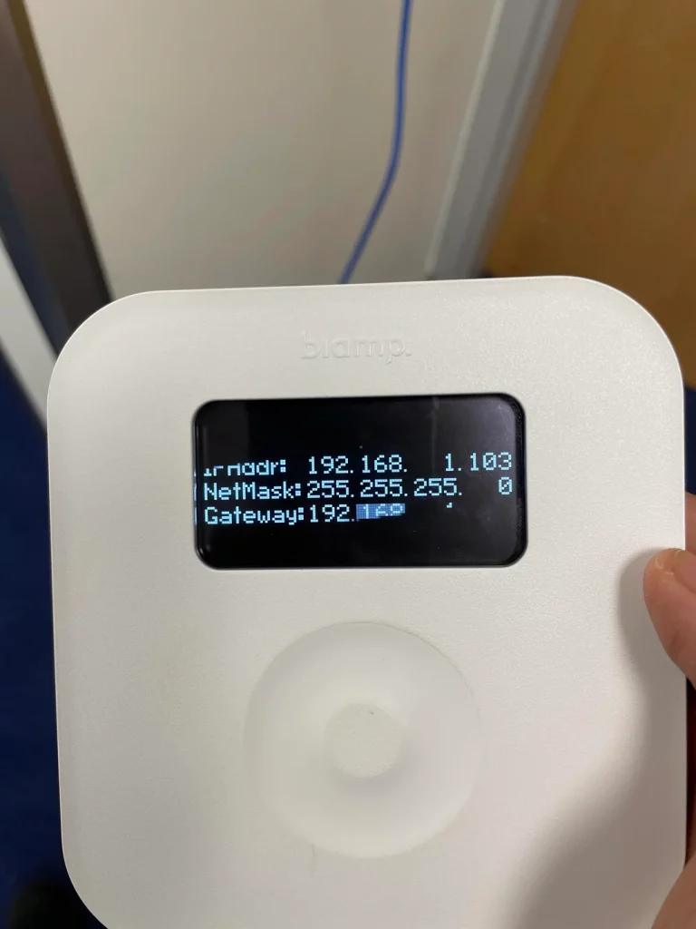 A close-up of a white Biamp Tec-1 control panel displaying network information on its black screen. The screen shows "IP Addr: 192.168.1.103", "NetMask: 255.255.255.0", and "Gateway: 192.168.1.1". Below the screen is a circular, textured control dial, and a thumb is partially visible on the right side of the device.