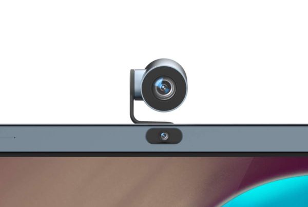 A black webcam is clipped to the top center of a laptop screen, with the laptop's built-in webcam visible directly below it. The screen displays a colorful, abstract background.