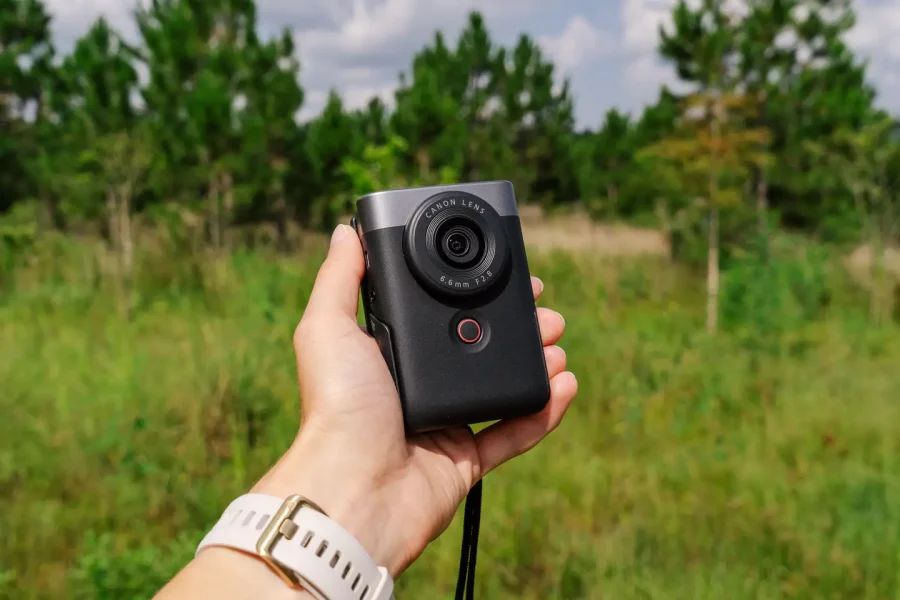 A hand holding the Canon PowerShot V10 Vlogging Camera outdoors, with a green forest and blue sky in the background.