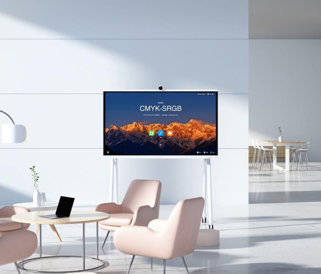 A white interactive display screen on a white mobile stand, centered in a modern, well-lit office or lounge area. The screen shows a vibrant mountain landscape with "CMYK-SRGB" text and app icons. In front of the screen, there's a light wooden coffee table with a black laptop on it, surrounded by two light pink armchairs. In the background to the right, a meeting area with a round table and white chairs is visible. The room features white walls and a minimalist design.