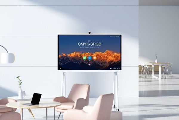 A white interactive display screen on a white mobile stand, centered in a modern, well-lit office or lounge area. The screen shows a vibrant mountain landscape with "CMYK-SRGB" text and app icons. In front of the screen, there's a light wooden coffee table with a black laptop on it, surrounded by two light pink armchairs. In the background to the right, a meeting area with a round table and white chairs is visible. The room features white walls and a minimalist design.