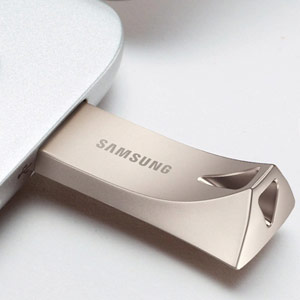 A close-up of a metallic Samsung Bar Plus 32GB USB flash drive, partially inserted into a silver laptop or device, showing the Samsung logo and the triangular loop design.