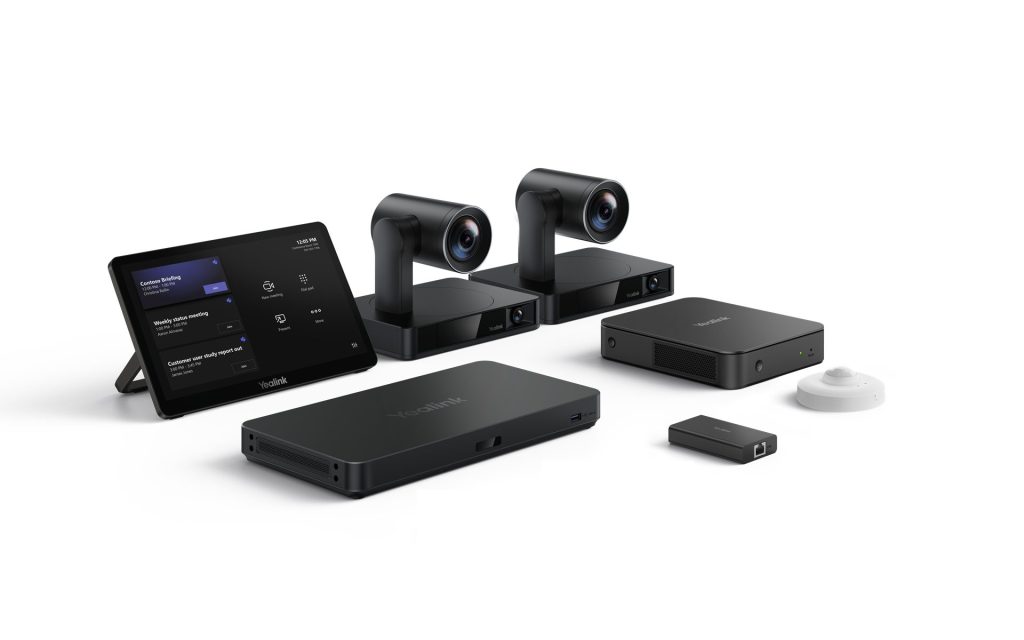 A professional setup of Yealink video conferencing equipment arranged on a white surface. From left to right, there's a black tablet displaying what appears to be a meeting schedule or interface. Behind it are two identical, sleek black cameras with cylindrical lenses mounted on rectangular bases. In front of the cameras are two rectangular black devices, one slightly larger than the other, both with the Yealink logo. To the far right, there's a small, rectangular black dongle and a small, white, circular device, likely a microphone or speaker.