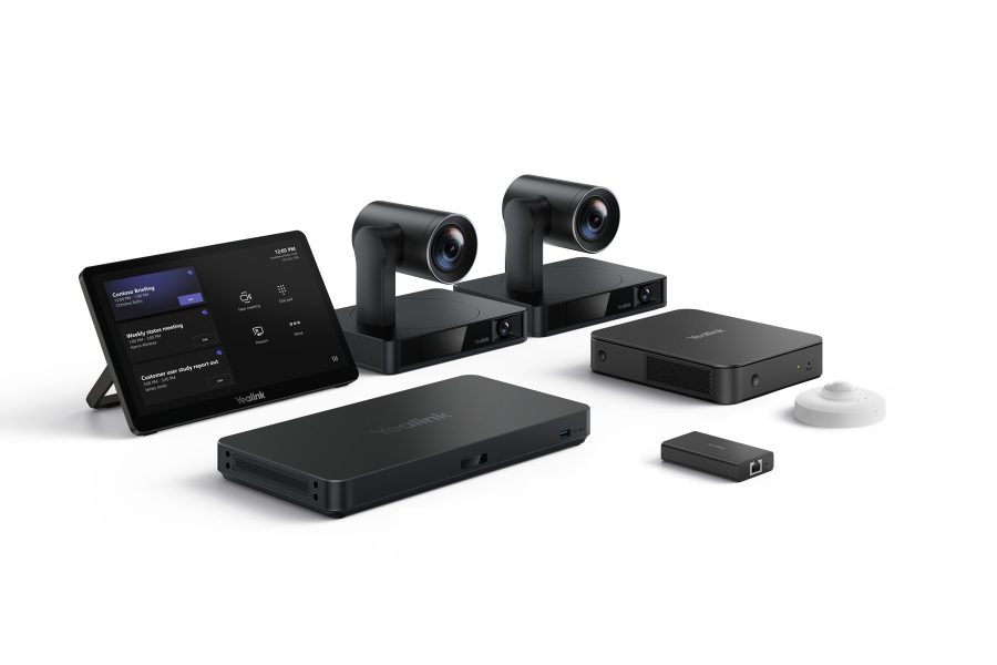 A professional setup of Yealink video conferencing equipment arranged on a white surface. From left to right, there's a black tablet displaying what appears to be a meeting schedule or interface. Behind it are two identical, sleek black cameras with cylindrical lenses mounted on rectangular bases. In front of the cameras are two rectangular black devices, one slightly larger than the other, both with the Yealink logo. To the far right, there's a small, rectangular black dongle and a small, white, circular device, likely a microphone or speaker.