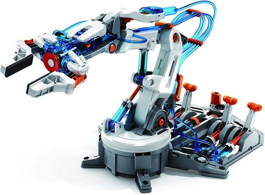 A close-up, three-quarter view shows a white and gray robotic arm kit with orange and translucent blue accents on a white background. The arm has a claw-like hand at its end. An array of orange levers and a series of pistons are visible at the base of the arm, connected to the arm itself by translucent blue tubing, suggesting it is a hydraulic or pneumatic toy.