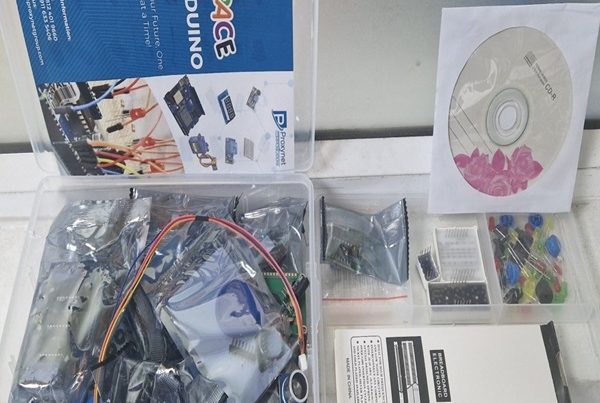 A transparent plastic box filled with various electronic components is shown on a light-colored surface. The box is open, revealing several smaller bags containing wires, circuit boards, and other parts, along with a white breadboard. To the left, a blue and white instruction booklet is visible, titled "iSPACE ARDUINO KIT." To the right of the main box, a smaller clear plastic container holds tiny colored components, and a white CD sleeve with a floral design containing a disc is also visible.