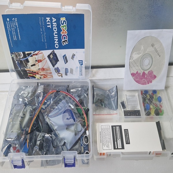 A transparent plastic box filled with various electronic components is shown on a light-colored surface. The box is open, revealing several smaller bags containing wires, circuit boards, and other parts, along with a white breadboard. To the left, a blue and white instruction booklet is visible, titled "iSPACE ARDUINO KIT." To the right of the main box, a smaller clear plastic container holds tiny colored components, and a white CD sleeve with a floral design containing a disc is also visible.