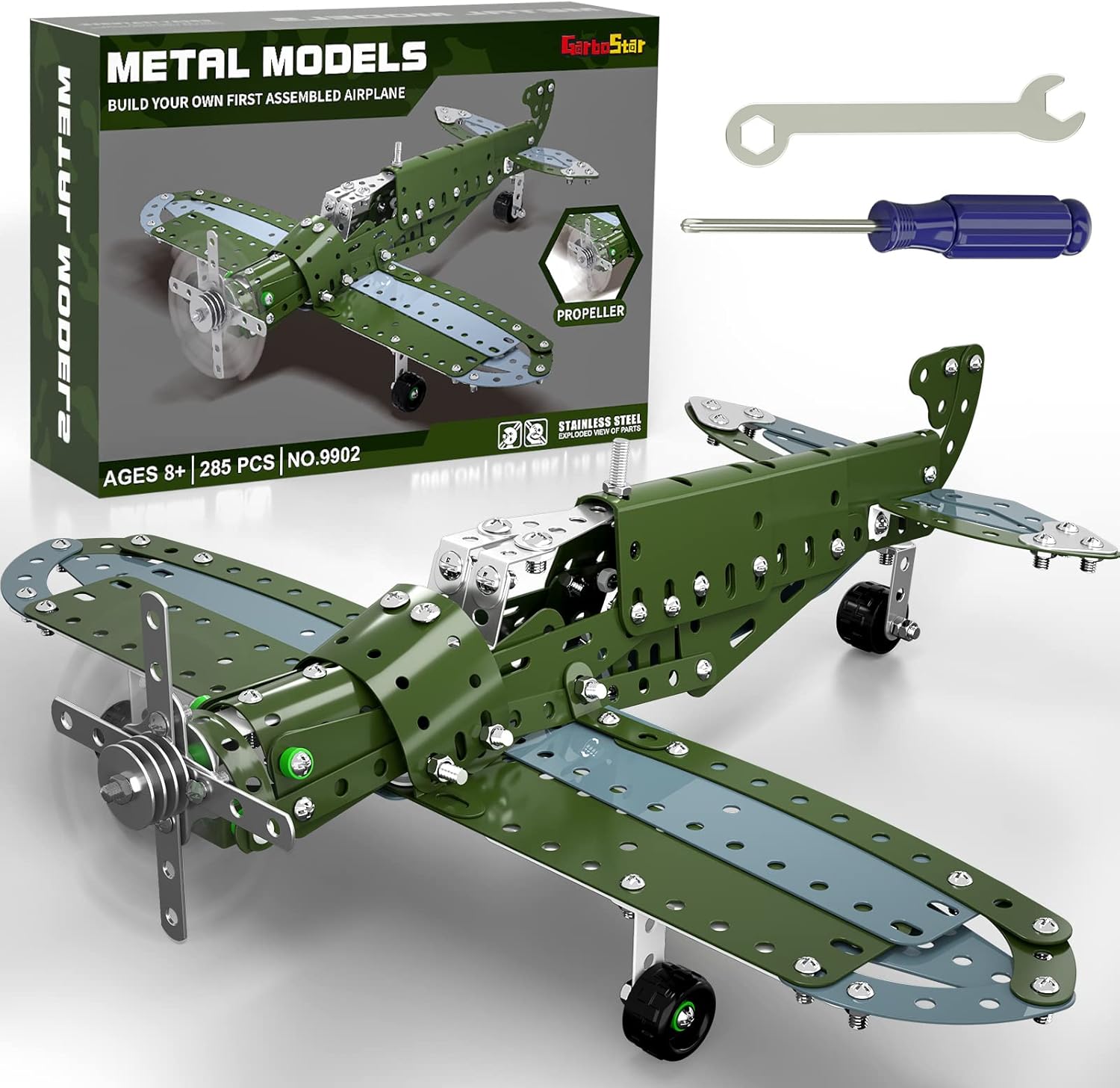 A metal construction toy kit of a green and gray airplane is displayed with its packaging and tools against a white background. The assembled airplane model, featuring an olive green fuselage and wings with light gray undersides and silver accents, is prominent in the foreground. A propeller is at the front, and black wheels are visible underneath. In the background, the kit's dark green box is shown, labeled "METAL MODELS" and "BUILD YOUR OWN FIRST ASSEMBLED AIRPLANE." The box also indicates "AGES 8+," "285 PCS," and "NO.9902," and mentions "STAINLESS STEEL." Above the box, a silver wrench and a blue-handled screwdriver, the tools used for assembly, are laid out.