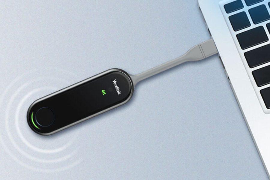 The image shows a black, oval-shaped device with the "Yealink" logo and "4K" text on it, connected via a short gray cable to a laptop's USB-C port. There's also a circular button on the left side of the device. The background appears to be a light-colored, possibly metallic, surface.