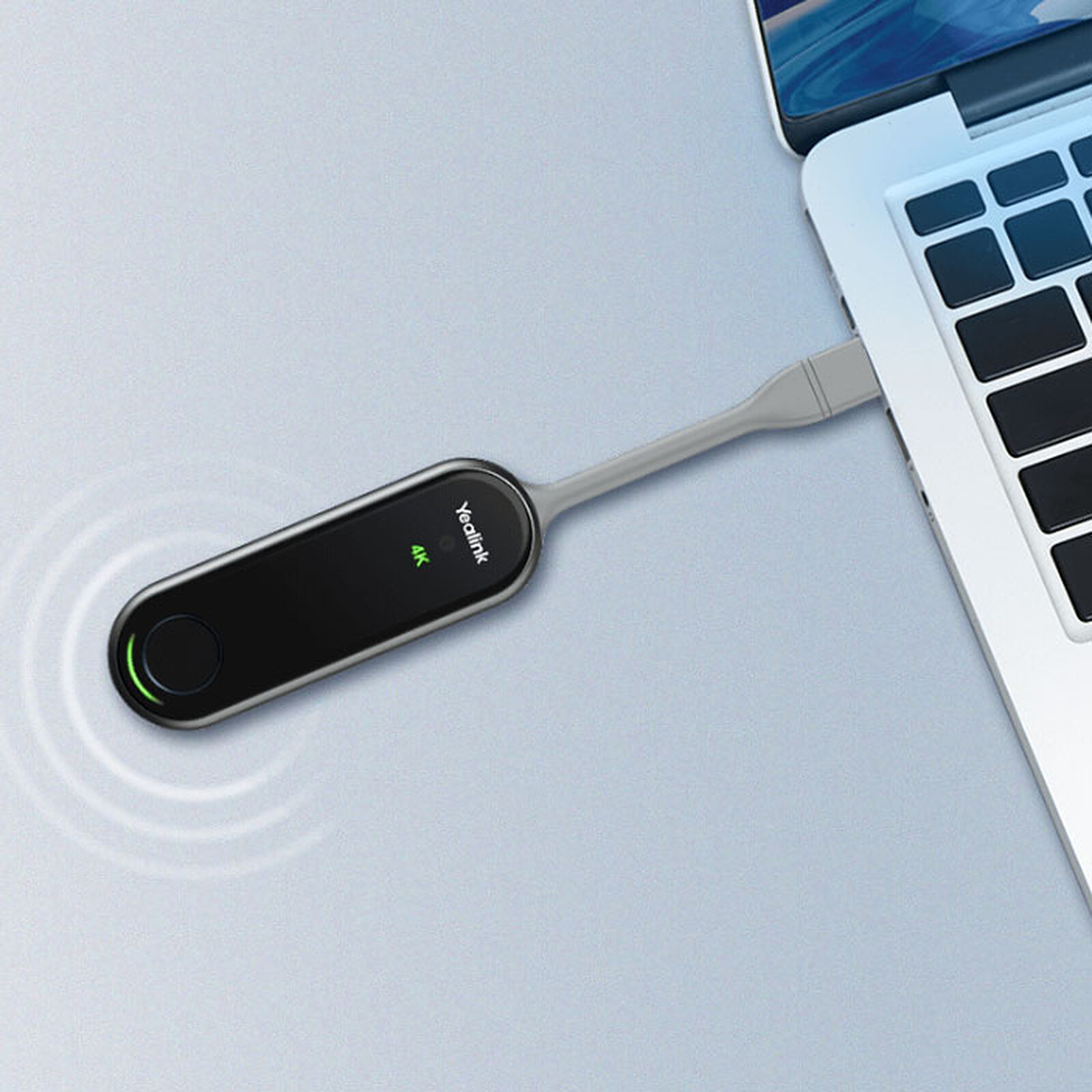 The image shows a black, oval-shaped device with the "Yealink" logo and "4K" text on it, connected via a short gray cable to a laptop's USB-C port. There's also a circular button on the left side of the device. The background appears to be a light-colored, possibly metallic, surface.