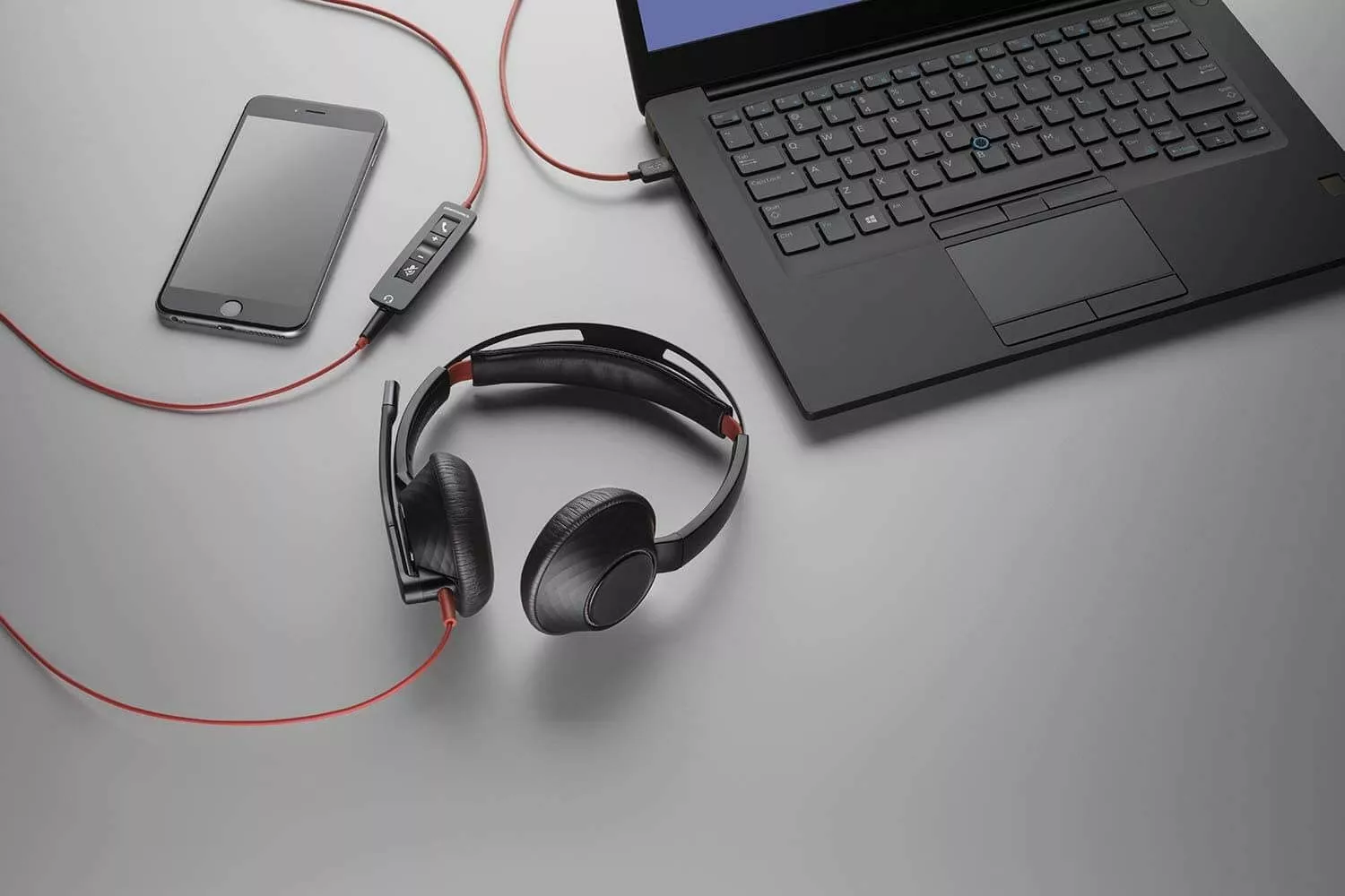 A flat lay, high-angle shot captures a black laptop, a smartphone, and a black headset with red accents on a gray surface. The laptop is positioned in the upper right, partially cropped, with a black cable extending from its right side. A black smartphone with a dark screen lies in the upper left. A black and red headset with an attached microphone boom is in the center foreground, its red cable snaking towards a small black inline control panel with buttons, which then connects to the laptop via a red cable. Another red cable from the control panel extends towards the smartphone.