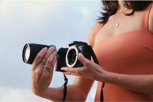 A cropped, mid-shot photo shows a person in an orange top holding a detached black camera lens in one hand and a black Canon EOS R10 camera body in the other. The camera body's sensor is visible. The person is wearing a gold necklace and several gold rings on their fingers. The background is a bright, slightly cloudy sky.