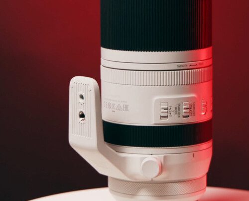 A side-angle, close-up shot of a white Canon RF 70-200mm f/2.8L IS USM lens. The lens is equipped with a tripod collar and rests on a white surface. The background is a dramatic red and black, highlighting the lens.