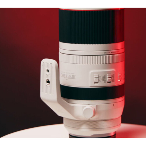 A side-angle, close-up shot of a white Canon RF 70-200mm f/2.8L IS USM lens. The lens is equipped with a tripod collar and rests on a white surface. The background is a dramatic red and black, highlighting the lens.