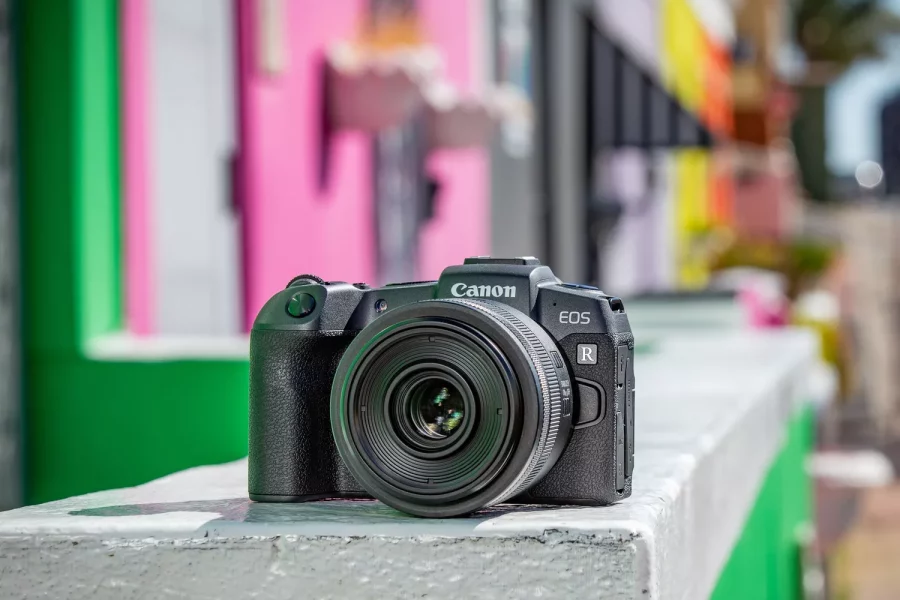 A high-quality product shot of a black Canon EOS RP mirrorless digital camera, viewed from a three-quarter angle on a white background. The camera's lens mount, body texture, and several top-panel controls are clearly visible.