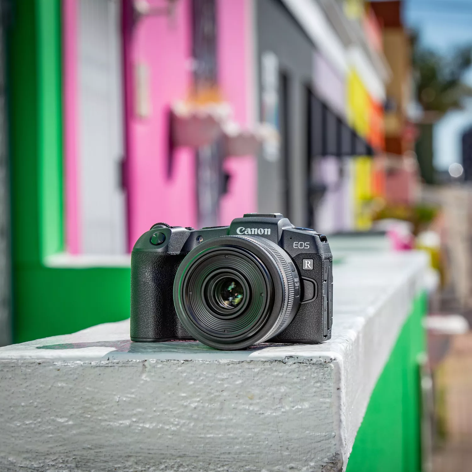 A high-quality product shot of a black Canon EOS RP mirrorless digital camera, viewed from a three-quarter angle on a white background. The camera's lens mount, body texture, and several top-panel controls are clearly visible.