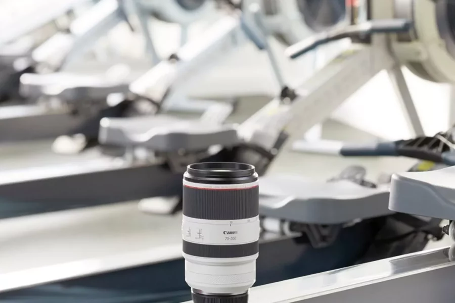A professional product shot of a Canon RF 70-200mm F2.8 L IS USM lens. The white lens is standing upright on the rail of a rowing machine in a gym, with more blurred rowing machines visible in the background.
