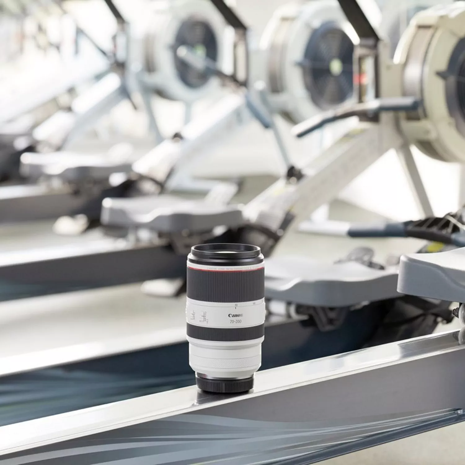 A professional product shot of a Canon RF 70-200mm F2.8 L IS USM lens. The white lens is standing upright on the rail of a rowing machine in a gym, with more blurred rowing machines visible in the background.