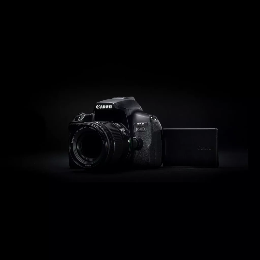 A dark, high-contrast, product-style studio shot of a black Canon EOS 80D camera. The camera is positioned at an angle, with a Canon lens attached and the LCD screen flipped out to the side. The lighting is focused on the camera body, creating a dramatic, dark background.