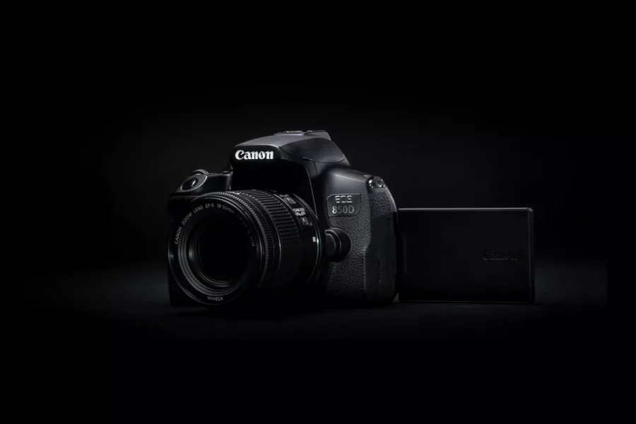 A dark, high-contrast, product-style studio shot of a black Canon EOS 80D camera. The camera is positioned at an angle, with a Canon lens attached and the LCD screen flipped out to the side. The lighting is focused on the camera body, creating a dramatic, dark background.