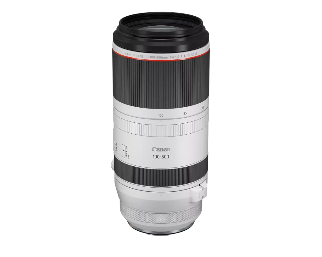 A product shot of a white Canon RF 100-500mm f/4.5-7.1L IS USM super-telephoto lens. The lens is isolated on a white background, standing upright with the lens hood mount at the top and the RF mount at the bottom. The zoom ring is black, while the rest of the lens barrel is white, with a red ring near the top.