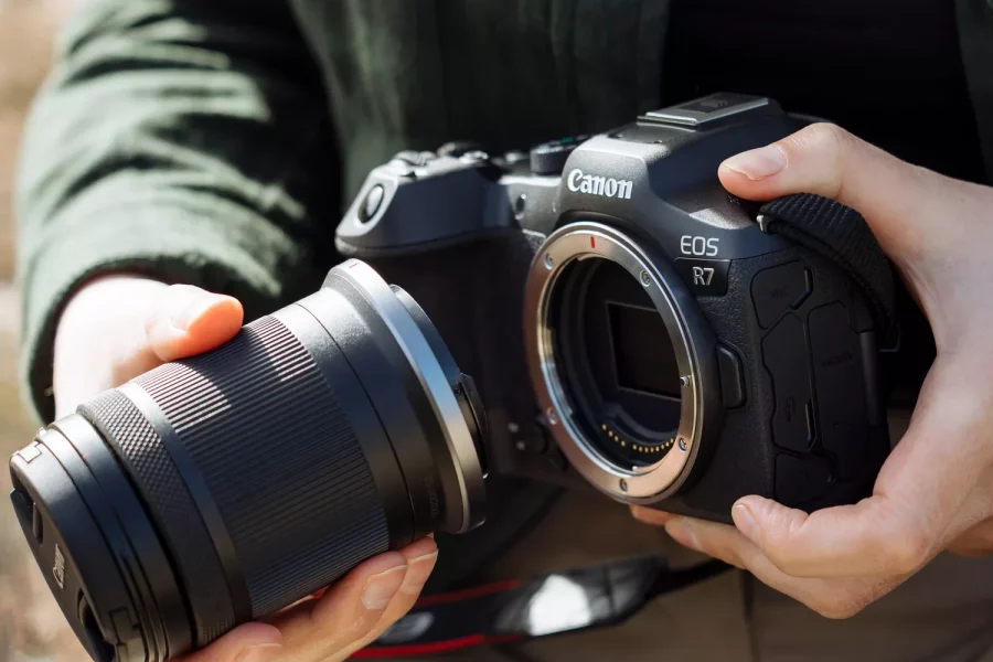 A person's hands are holding a black Canon EOS R7 mirrorless camera with the lens detached. The lens, a black Canon RF-S 18-150mm, is held in their left hand, ready to be mounted onto the camera body.