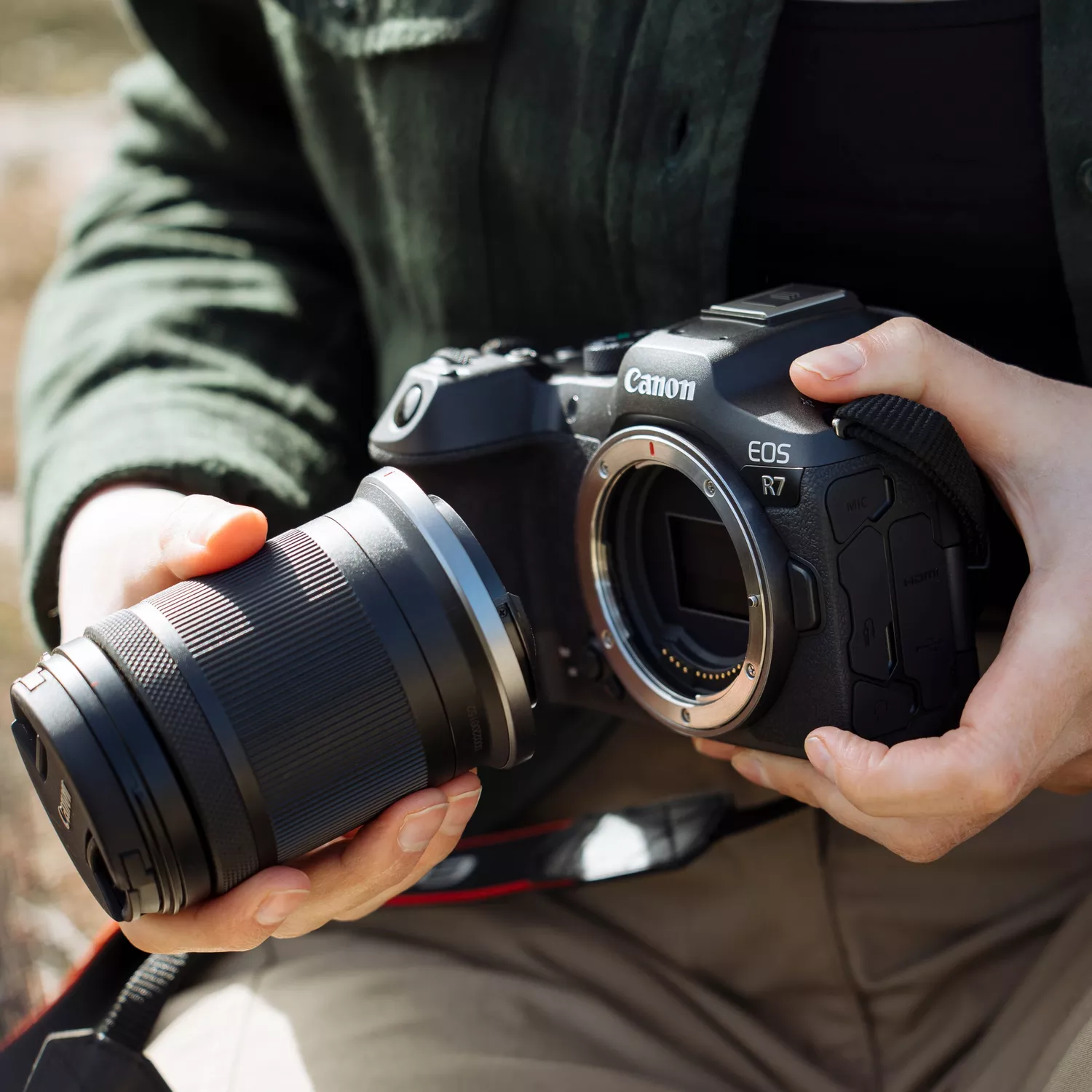 A person's hands are holding a black Canon EOS R7 mirrorless camera with the lens detached. The lens, a black Canon RF-S 18-150mm, is held in their left hand, ready to be mounted onto the camera body.