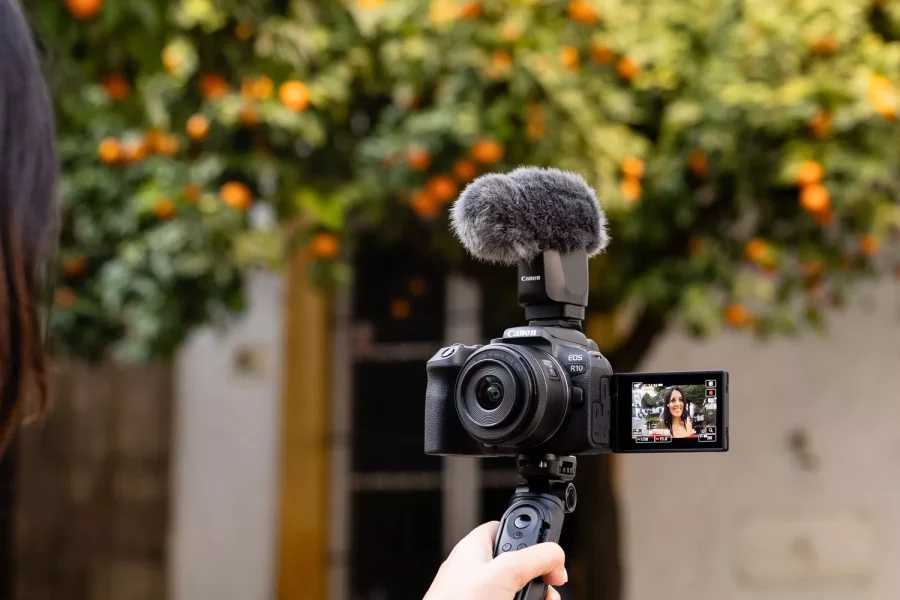 A person is holding a Canon EOS R10 mirrorless camera with an RF-S 18-150mm lens and a microphone attached. The camera's screen is flipped out, showing a self-portrait view. The background is out of focus, featuring an orange tree and a building.