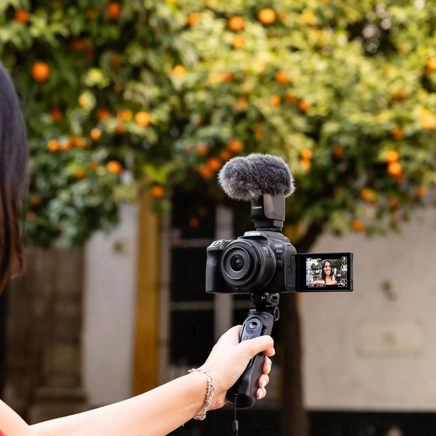A person is holding a Canon EOS R10 mirrorless camera with an RF-S 18-150mm lens and a microphone attached. The camera's screen is flipped out, showing a self-portrait view. The background is out of focus, featuring an orange tree and a building.