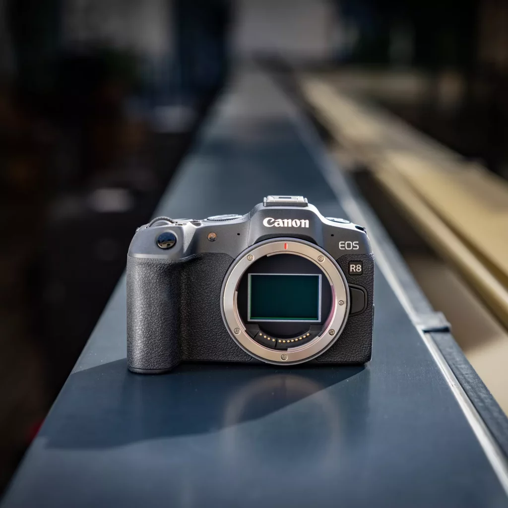 A black Canon EOS R8 camera body without a lens sits on a dark-colored railing. The camera is facing forward, with its lens mount visible and its sensor reflecting light. The background is a blurred outdoor urban scene, creating a shallow depth of field.