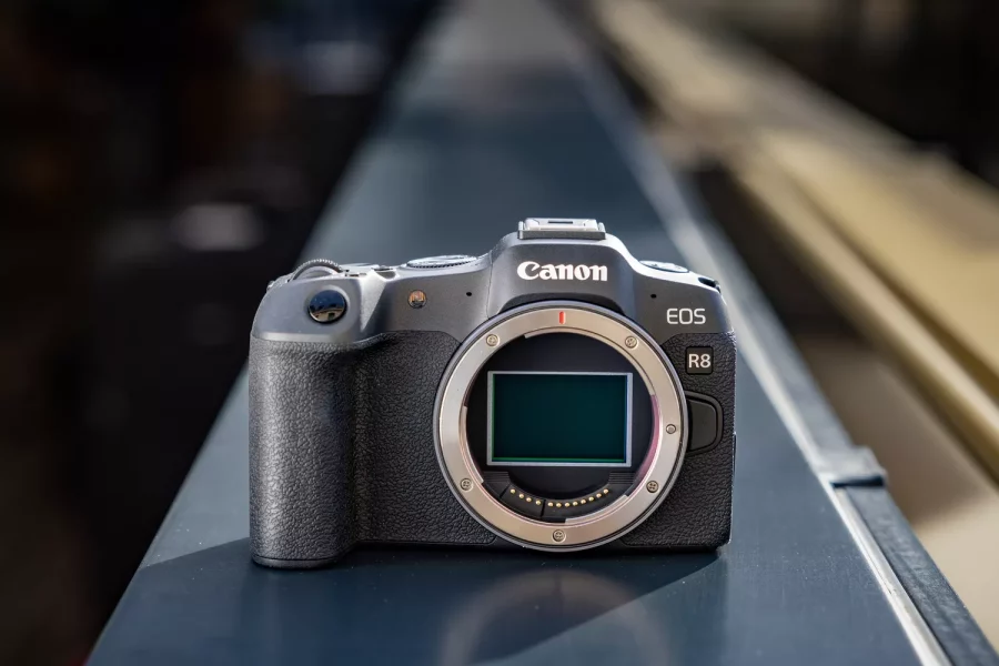 A black Canon EOS R8 camera body without a lens sits on a dark-colored railing. The camera is facing forward, with its lens mount visible and its sensor reflecting light. The background is a blurred outdoor urban scene, creating a shallow depth of field.