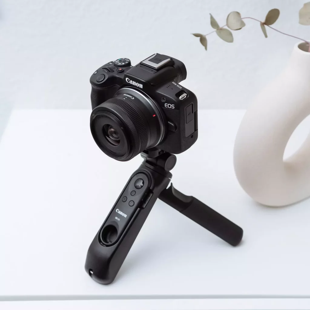 A three-quarters view of a black Canon EOS R50 camera mounted on a Canon HG-100TBR tripod grip. The camera and tripod are sitting on a white surface, with a minimal white vase and a branch in the background.