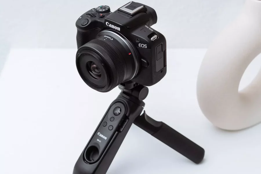 A three-quarters view of a black Canon EOS R50 camera mounted on a Canon HG-100TBR tripod grip. The camera and tripod are sitting on a white surface, with a minimal white vase and a branch in the background.