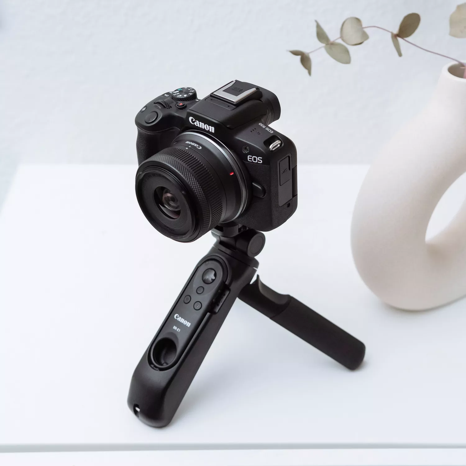 A three-quarters view of a black Canon EOS R50 camera mounted on a Canon HG-100TBR tripod grip. The camera and tripod are sitting on a white surface, with a minimal white vase and a branch in the background.