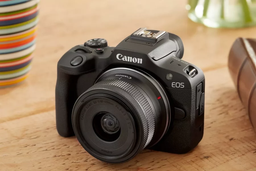 Based on the image, the camera is a **Canon EOS R100**. The lens attached to it is a **Canon RF-S 18-45mm f/4.5-6.3 IS STM**. The camera is resting on a wooden surface, with a colorful cup on the left and a glass vase with a plant on the right.