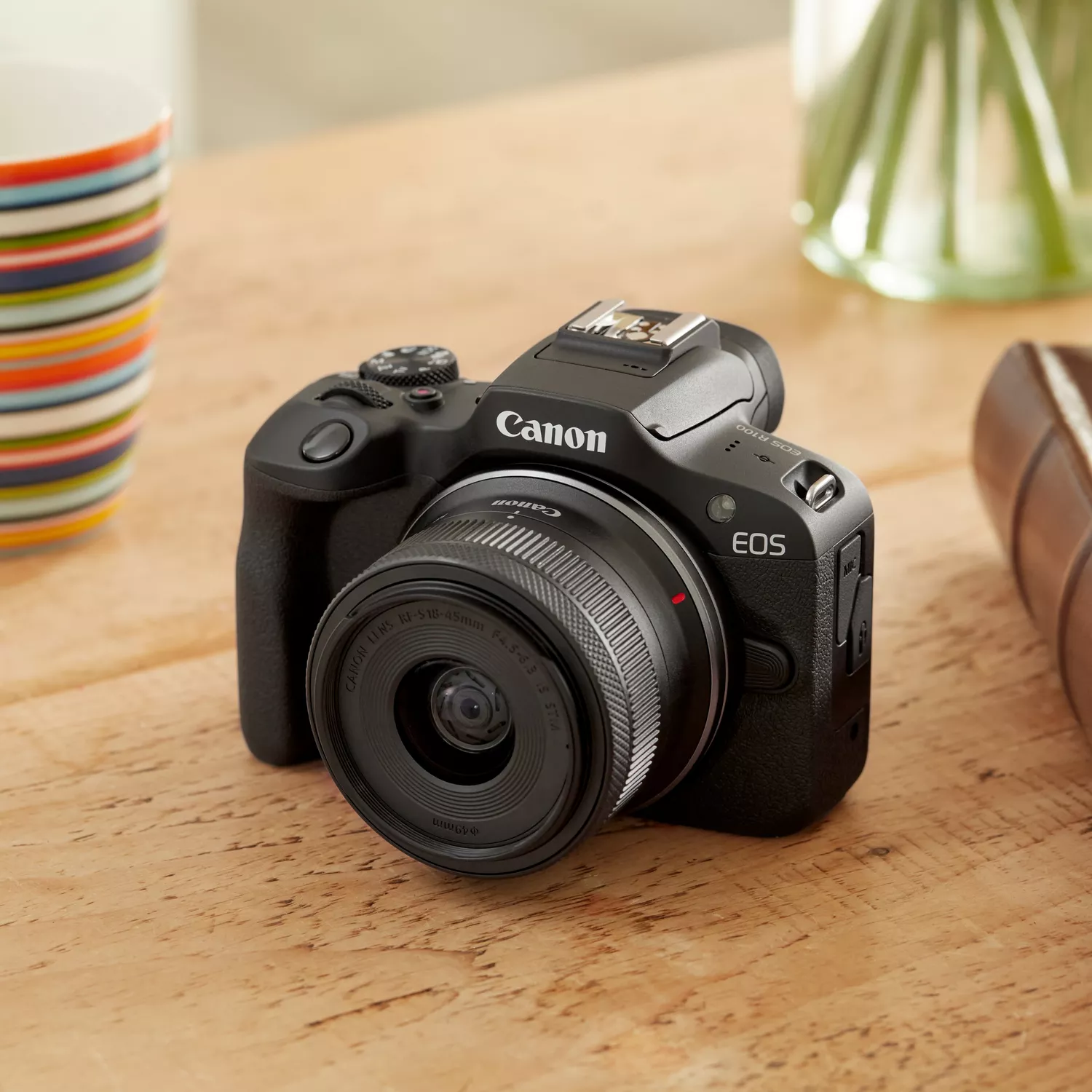 Based on the image, the camera is a **Canon EOS R100**. The lens attached to it is a **Canon RF-S 18-45mm f/4.5-6.3 IS STM**. The camera is resting on a wooden surface, with a colorful cup on the left and a glass vase with a plant on the right.