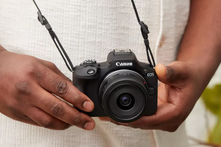 A close-up, eye-level shot of a person's hands holding a black Canon EOS R100 camera with a lens attached, the camera is hanging from a black neck strap and the person is wearing a white collared shirt.