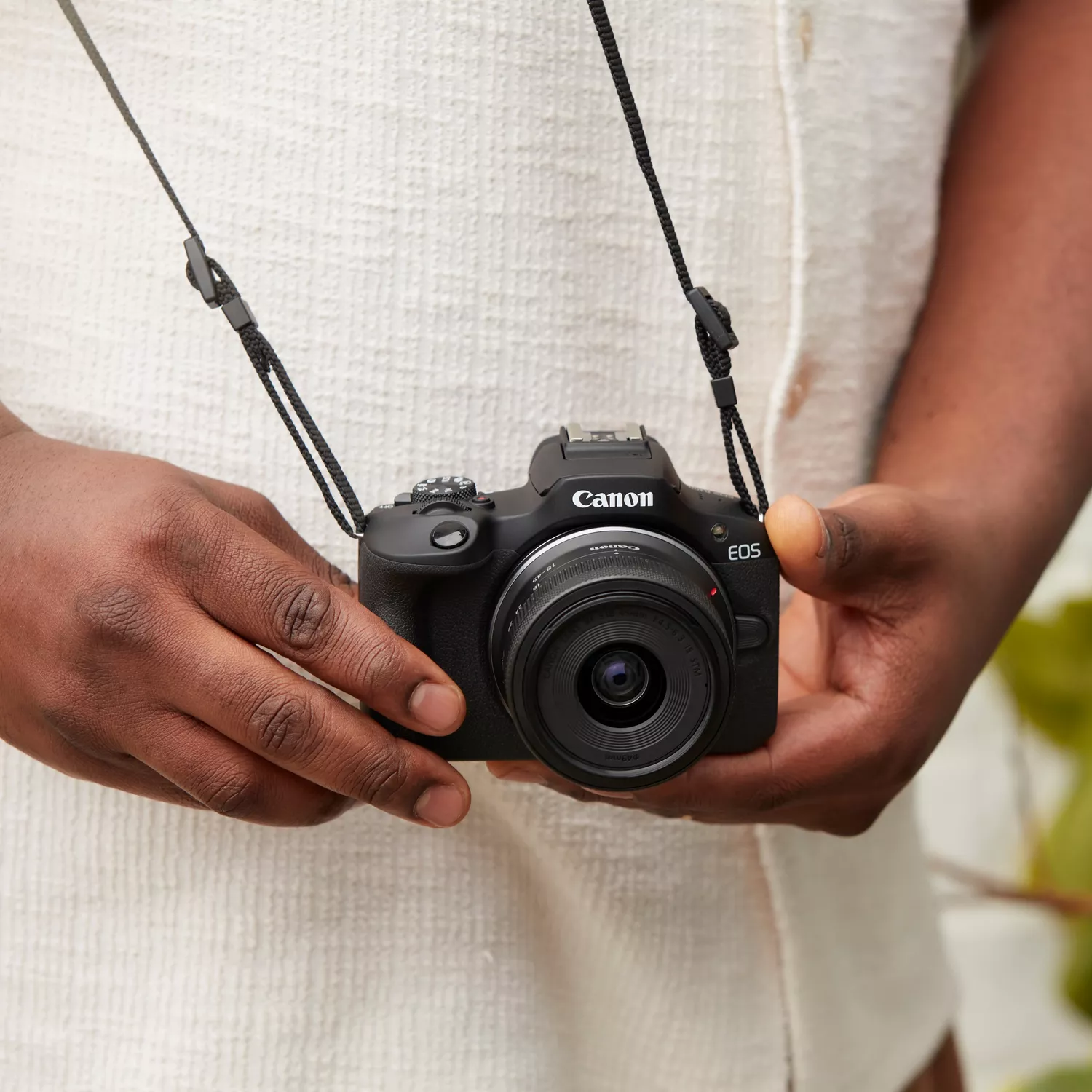 A close-up, eye-level shot of a person's hands holding a black Canon EOS R100 camera with a lens attached, the camera is hanging from a black neck strap and the person is wearing a white collared shirt.