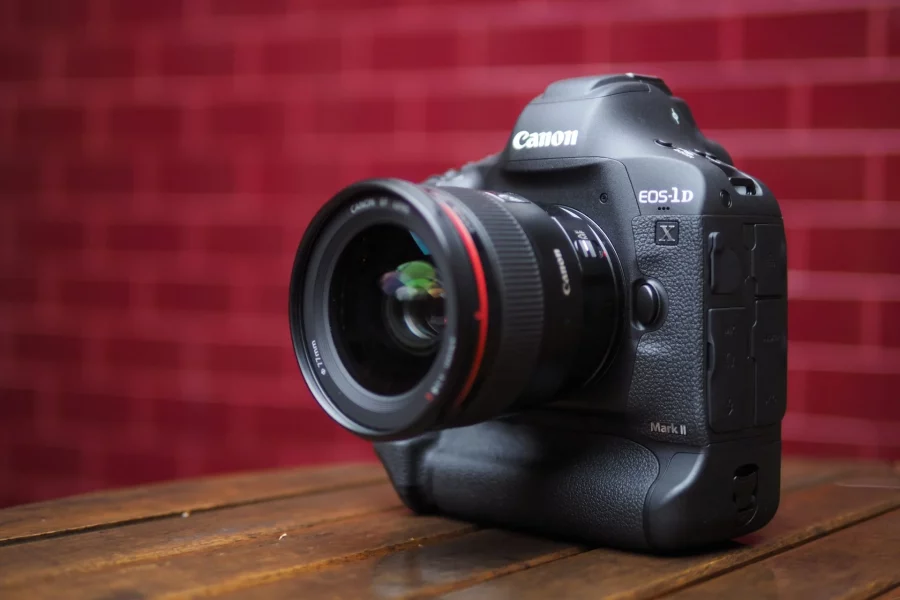 A low-angle shot of a black Canon EOS-1D X Mark II DSLR camera with a lens attached, sitting on a wooden table. The camera is positioned at an angle, showing its front and side. The background is a blurry red brick wall.