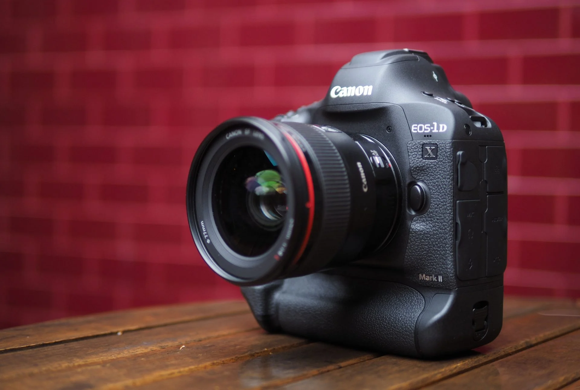 A low-angle shot of a black Canon EOS-1D X Mark II DSLR camera with a lens attached, sitting on a wooden table. The camera is positioned at an angle, showing its front and side. The background is a blurry red brick wall.