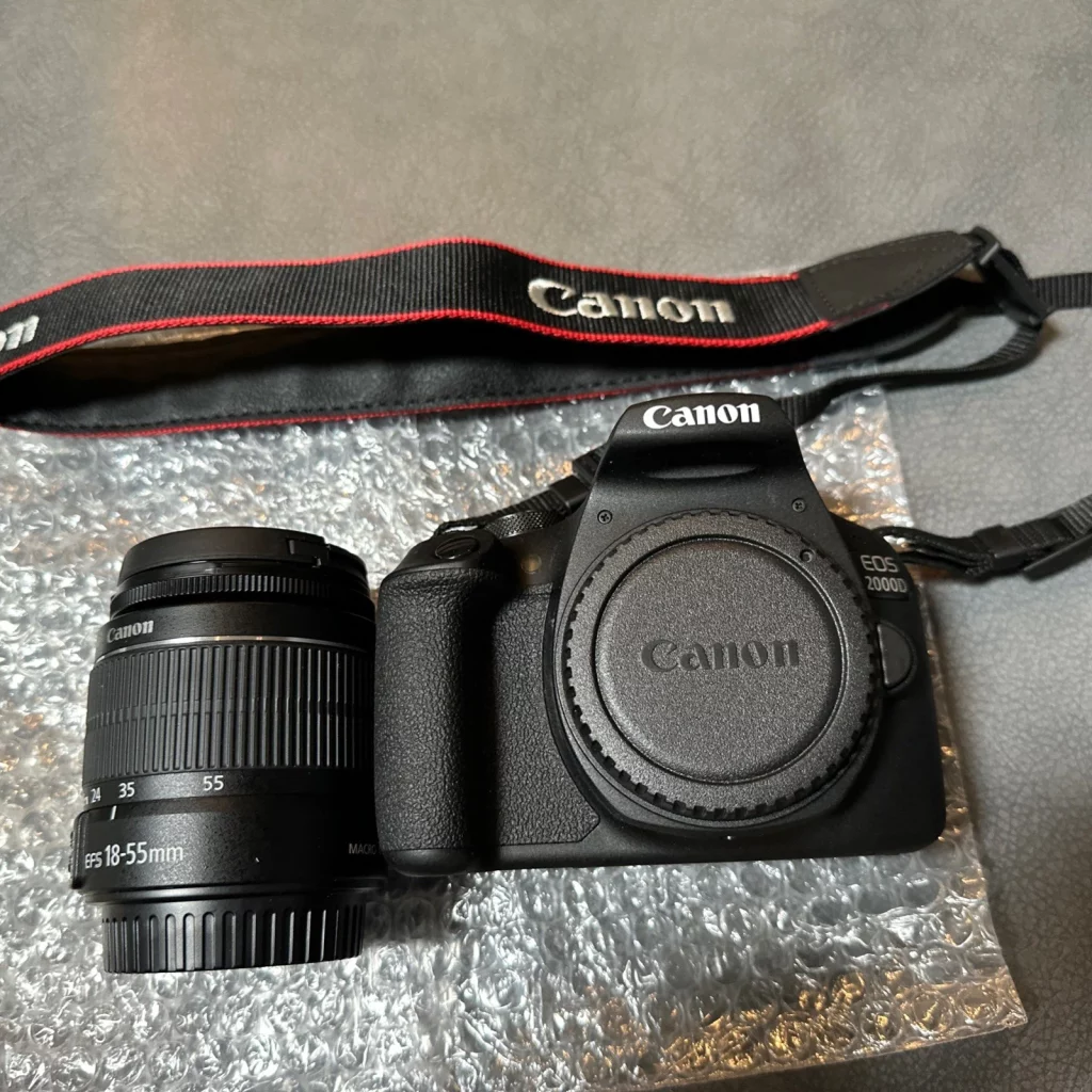 A flat lay shot of a Canon EOS 2000D camera kit on a gray surface with bubble wrap. The kit includes a black Canon EOS 2000D camera body with a lens cap on, a separate Canon EF-S 18-55mm lens, and a black and red Canon camera strap.